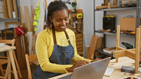 Téléchargez les photos : Femme afro-américaine utilisant un ordinateur portable dans un atelier de menuiserie entouré d'outils de travail du bois - en image libre de droit