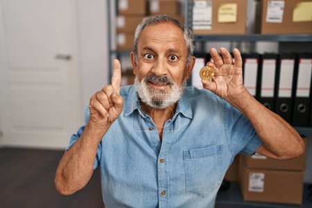 Téléchargez les photos : Homme âgé joyeux au bureau, pointant du doigt avec confiance avec le visage ravi, tenant Bitcoin pièce, tous allumés avec une idée intelligente et une question ahurissante! - en image libre de droit