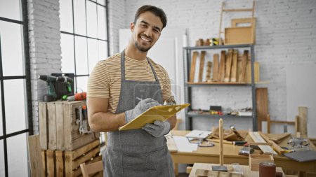 Photo for A handsome young hispanic man with a beard smiling in a carpentry workshop, wearing an apron and holding a clipboard. - Royalty Free Image