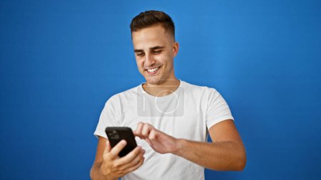 Photo for A cheerful young hispanic man in a white shirt using a smartphone against a blue background - Royalty Free Image