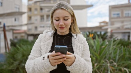 A young blonde woman in a cozy jacket uses her smartphone, surrounded by urban greenery.