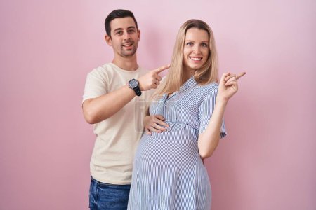 Téléchargez les photos : Jeune couple s'attendant à un bébé debout sur fond rose avec un grand sourire sur le visage, pointant du doigt vers le côté en regardant la caméra. - en image libre de droit
