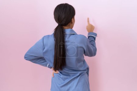 Foto de Mujer árabe joven con pijama azul posando hacia atrás apuntando hacia delante con la mano del dedo - Imagen libre de derechos