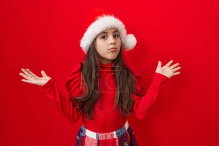 Photo for Adorable hispanic girl in christmas hat on isolated red background, shrugging and wearing confused expression  a clueless 'i don't know' gesture - Royalty Free Image