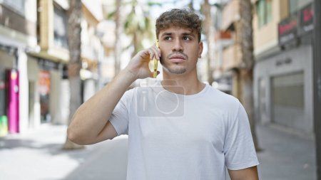 Photo for Cool-looking, handsome young hispanic man seriously engaged in conversation on his smartphone, standing under the sunny urban street's casual fashion backdrop with a concentrated expression. - Royalty Free Image