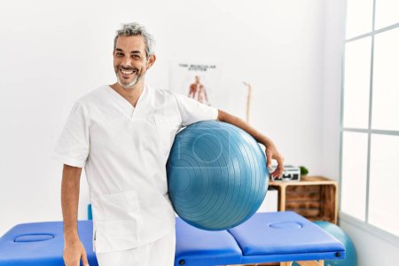 Téléchargez les photos : Homme moyen aux cheveux gris physiothérapeute souriant confiant tenant tapis de yoga à la clinique de réadaptation - en image libre de droit