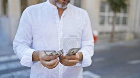 Schöner junger hispanischer Mann mit grauen Haaren, lässig auf einer sonnigen Straße der Stadt stehend, leidenschaftlich amerikanische Dollars zählend, ein Blick auf städtischen Reichtum und Investitionen.
