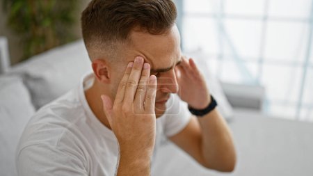 Un joven estresado en casa tocándose la frente e ilustrando un dolor de cabeza o preocupación con un fondo borroso.