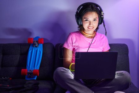 Young hispanic woman using laptop and headphones sitting on sofa at home
