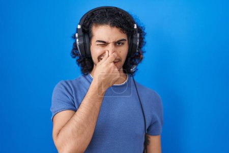 Téléchargez les photos : Homme hispanique aux cheveux bouclés écoutant de la musique à l'aide d'écouteurs sentant quelque chose de puant et dégoûtant, odeur intolérable, retenant son souffle avec les doigts sur le nez. mauvaise odeur - en image libre de droit