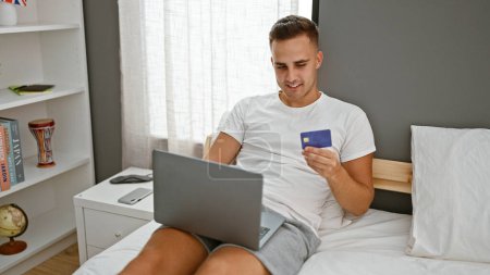 Junger hispanischer Mann im Schlafzimmer mit Laptop und Kreditkarte, der Online-Shopping oder Rechnungszahlung darstellt.