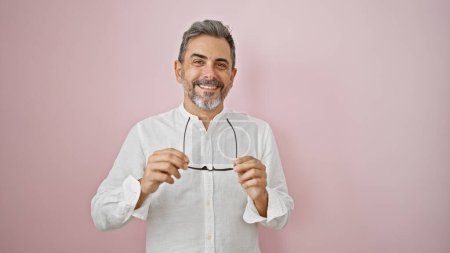 Foto de Joven hombre hispano confiado con el pelo gris, riendo alegremente mientras sostiene las gafas, de pie casualmente sobre un fondo rosa aislado, exudando felicidad y estilo de vida fresco - Imagen libre de derechos