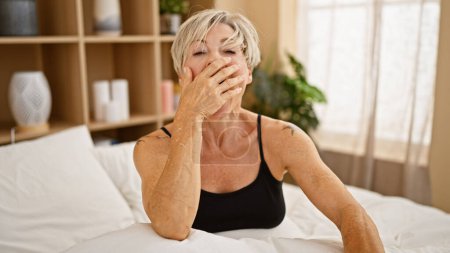 Foto de Una mujer madura con el pelo gris corto parece cansada mientras está sentada en un dormitorio con un interior acogedor. - Imagen libre de derechos