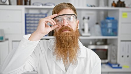 Photo for Relaxed yet serious-faced, young redhead male scientist, amped about a groundbreaking experiment in the lab, ensures safety with security glasses - Royalty Free Image