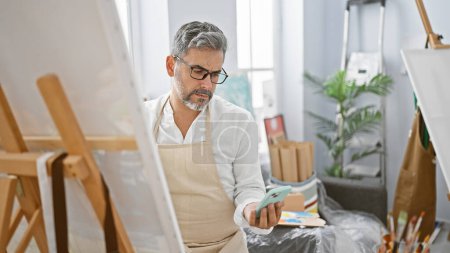 Téléchargez les photos : Jeune homme hispanique attrayant, artiste aux cheveux gris, absorbé par le dessin sur écran de smartphone à son atelier d'art confortable. - en image libre de droit