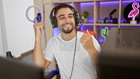 A smiling young man with a beard enjoying gaming in a well-lit room at night, wearing headphones and giving a thumbs-up.
