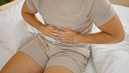 Foto de Guapo joven hispano sufriendo en agonía con dolor de estómago, sentado en su cama tocando su abdomen, expresión infeliz en su dormitorio hogareño - Imagen libre de derechos