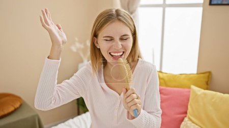 Photo for A joyful woman singing using a hairbrush as a microphone in a cozy, well-lit bedroom setting. - Royalty Free Image
