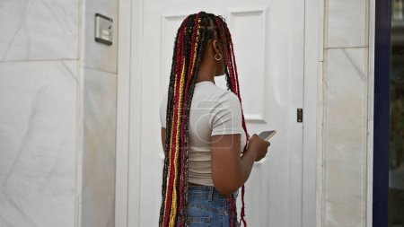 Foto de ¡Trenzas geniales! impresionante mujer afroamericana negra seriamente concentrándose en su teléfono inteligente, de pie en vibrante estilo de vida de la calle de la ciudad soleada. - Imagen libre de derechos