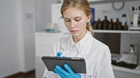 Eine blonde Frau im Laborkittel konzentriert sich auf eine Tablette in einem wissenschaftlichen Labor, umgeben von Geräten.