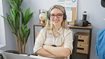 Téléchargez les photos : Une jeune femme caucasienne souriante portant écouteurs et lunettes, bras croisés, se tient dans un bureau moderne. - en image libre de droit