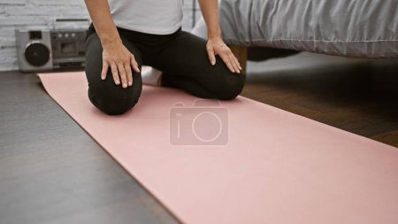 Téléchargez les photos : Une femme calme assise sur un tapis de yoga dans la chambre, les mains reposant sur les genoux, expérimentant la relaxation matinale, l'équilibre et la concentration grâce à l'exercice méditatif - en image libre de droit