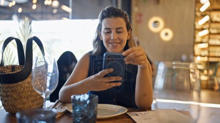 Téléchargez les photos : Jeune belle femme hispanique souriant heureux en utilisant smartphone assis sur la table au restaurant - en image libre de droit