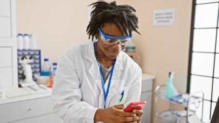 Foto de Mujer afroamericana con rastas, vistiendo bata de laboratorio y gafas, usando teléfono inteligente en un entorno de laboratorio. - Imagen libre de derechos