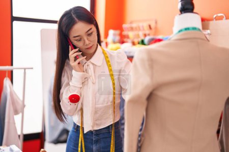 Téléchargez les photos : Jeune femme chinoise tailleur parler sur smartphone regarder veste à l'atelier - en image libre de droit