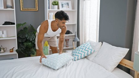 Ein junger hispanischer Mann arrangiert Kissen in einem modernen Schlafzimmer zu Hause und präsentiert ein aufgeräumtes und stilvolles Interieur.