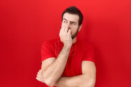 Foto de Hombre hispano joven con camiseta roja casual que parece estresado y nervioso con las manos en la boca mordiendo las uñas. problema de ansiedad. - Imagen libre de derechos