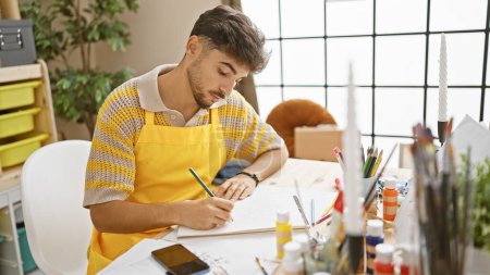 Photo for Handsome young arab man, a dedicated artist, fully absorbed in drawing on his notebook in a vibrant art studio - Royalty Free Image