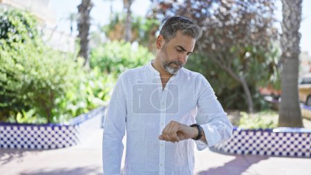 Worried young hispanic man with grey hair anxiously checks time on his wristwatch in the city park. a handsome face reveals serious concern of possibly being late.