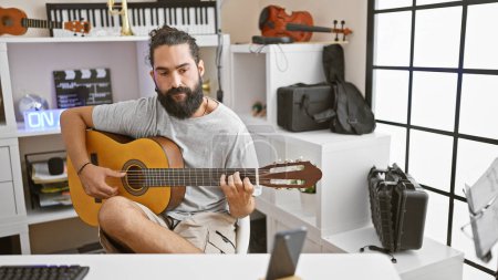 Handsome hispanic man plays acoustic guitar in a well-equipped modern music studio.