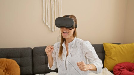 Téléchargez les photos : Une jeune femme joyeuse profitant d'un casque de réalité virtuelle dans un salon confortable, exprimant l'excitation et l'aventure dans un environnement domestique moderne. - en image libre de droit