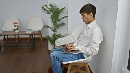 Photo for Handsome young hispanic man engrossed in serious business on laptop, sitting in chair at bustling waiting room - Royalty Free Image