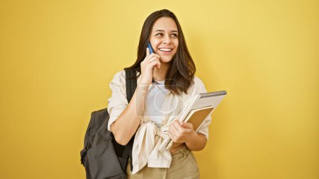 Photo for Confident young hispanic woman student, joyfully talking on phone, standing isolated on yellow background, juggling books smartly, displaying her casual, fun lifestyle with a radiant smile. - Royalty Free Image