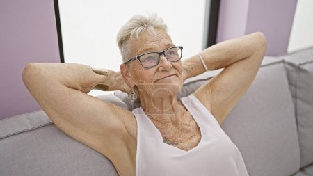 Téléchargez les photos : Femme âgée confiante aux cheveux gris, souriante et détendue, assise dans un salon confortable à la maison. jouissant d'un moment de bonheur, les mains reposant sur sa tête. - en image libre de droit