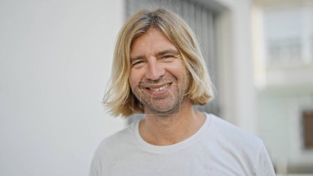 Photo for A smiling man with long blond hair posing outdoors on a sunny urban street. - Royalty Free Image