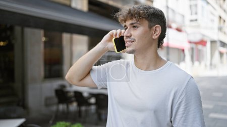 Téléchargez les photos : Jeune homme hispanique confiant profitant de la vie citadine décontractée, souriant, parlant sur smartphone et se baignant au soleil sur la terrasse du café - en image libre de droit