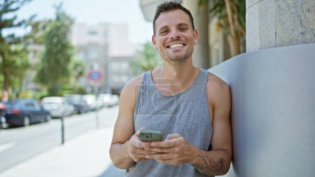 Foto de Sonriente joven hispano con barba y tatuaje se apoya en una esquina urbana mientras usa un teléfono inteligente - Imagen libre de derechos