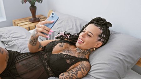 Téléchargez les photos : Confiant hispanique amputé femme textos sur smartphone, à la recherche sexy dans la lumière du matin, couché dans la chambre - en image libre de droit