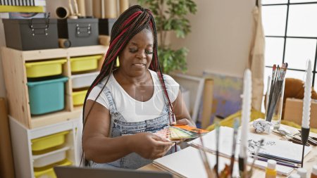 Inside the art studio, beautiful african american woman artist with braids confidently choosing color on laptop while immersed in her painting hobby
