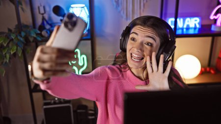 Photo for Young hispanic woman streaming videogames in a dark room, wearing headphones, waving at webcam, neon lights background. - Royalty Free Image