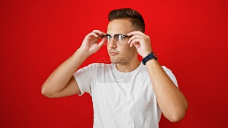 Foto de Guapo joven hispano ajustando gafas sobre fondo rojo aislado, retratando la confianza - Imagen libre de derechos