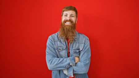 Foto de Joven chico alegre irlandés con el pelo rojo, sonriendo, barba, y brazos cruzados de pie solo sobre fondo rojo expresando vibraciones positivas - Imagen libre de derechos
