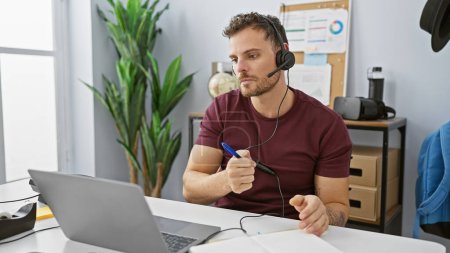 Photo for A focused hispanic man wearing a headset working indoors at an office with a laptop and notepad. - Royalty Free Image