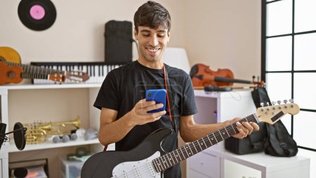 Téléchargez les photos : Jeune hispanique séduisant, musicien de guitare électrique, souriant avec charme en textant sur son smartphone dans un studio de musique, créant son propre concert de rock - en image libre de droit