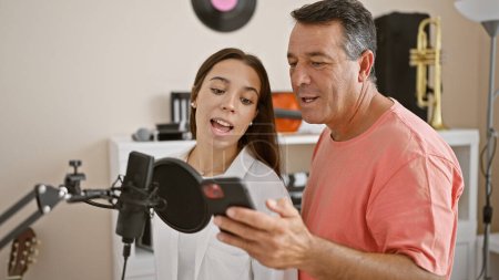 Photo for Confident man and woman musicians singing melody together, reading the song from a smartphone screen in a music studio, smiling as they create acoustic sound - Royalty Free Image