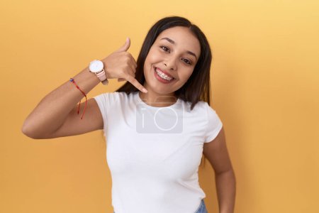 Téléchargez les photos : Jeune femme arabe portant t-shirt blanc décontracté sur fond jaune souriant faire geste de téléphone avec la main et les doigts comme parler au téléphone. communiquer les concepts. - en image libre de droit